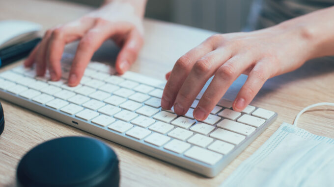 Close up of hands of a woman typing on a modern wireless keyboard. Medical mask by its side stock photo